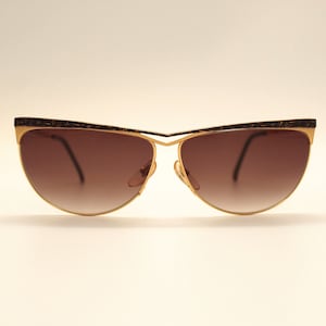Puede incluir: Un par de gafas de sol doradas con lentes de degradado marrón. Las gafas tienen forma de ojo de gato y un fino marco de metal.