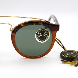 May include: Tortoise shell Ray-Ban sunglasses with gold accents and green lenses. The sunglasses have a round shape and a Ray-Ban logo on the right lens. The sunglasses are accompanied by a black Ray-Ban tag with gold lettering.