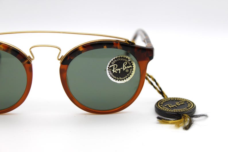May include: Tortoise shell Ray-Ban sunglasses with gold accents and green lenses. The sunglasses have a round shape and a Ray-Ban logo on the right lens. The sunglasses are accompanied by a black Ray-Ban tag with gold lettering.