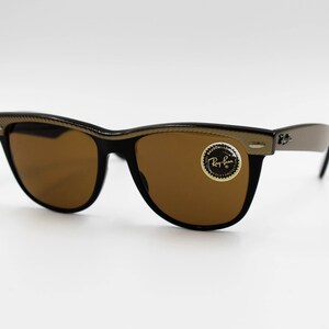 May include: Black sunglasses with brown lenses and a gold-colored frame. The sunglasses have a Ray-Ban logo on the right lens.