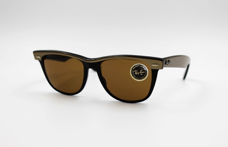 May include: Black sunglasses with brown lenses and a gold-colored frame. The sunglasses have a Ray-Ban logo on the right lens.