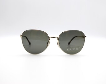 Puede incluir: Gafas de sol con montura dorada y lentes oscuras. Las monturas redondas tienen un diseño clásico. El nombre de la marca "GUCCI" está impreso en la lente derecha. Las gafas están sobre un fondo blanco.