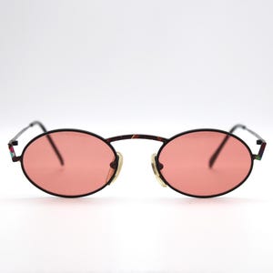 May include: Oval-shaped sunglasses with a black frame and pink lenses. The bridge and temples have a tortoiseshell pattern. The arms have a colorful design at the ends. A stylish accessory for eye protection.