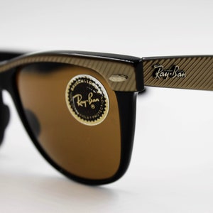 May include: Black Ray-Ban sunglasses with brown lenses and a gold-toned, textured frame. The sunglasses have a Ray-Ban logo on the right arm.