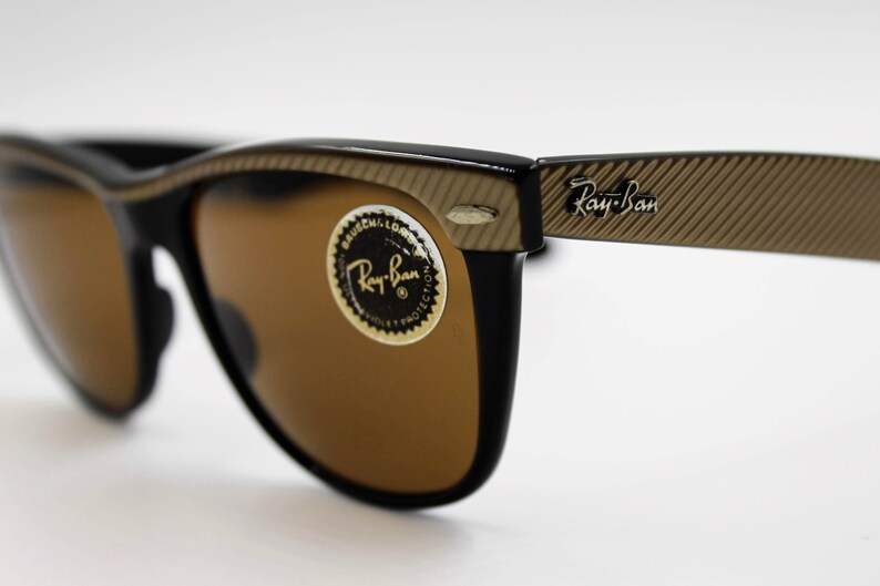 May include: Black Ray-Ban sunglasses with brown lenses and a gold-toned, textured frame. The sunglasses have a Ray-Ban logo on the right arm.