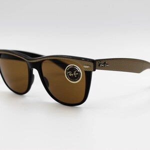 May include: Black Ray-Ban sunglasses with brown lenses and a gold-tone frame. The frame has a textured pattern and the Ray-Ban logo is visible on the right arm.