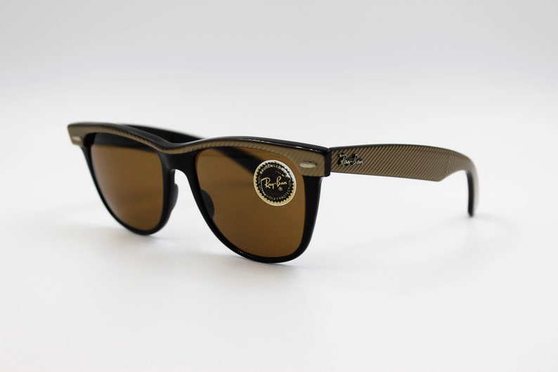 May include: Black Ray-Ban sunglasses with brown lenses and a gold-tone frame. The frame has a textured pattern and the Ray-Ban logo is visible on the right arm.