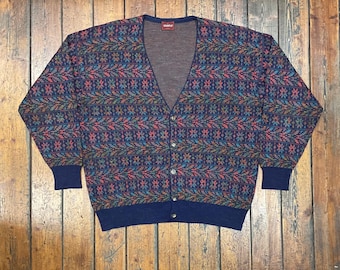 May include: A vintage cardigan sweater with a V-neck and button closure. The sweater features a repeating pattern of colorful geometric shapes in shades of blue, red, green, and brown. The cuffs and hem are solid navy blue.