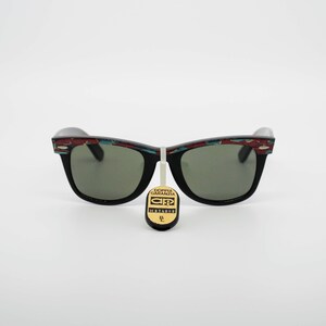 May include: Black sunglasses with green lenses and a decorative pattern on the top of the frames. The pattern includes red, blue, and white geometric shapes. A tag with text hangs from the sunglasses.