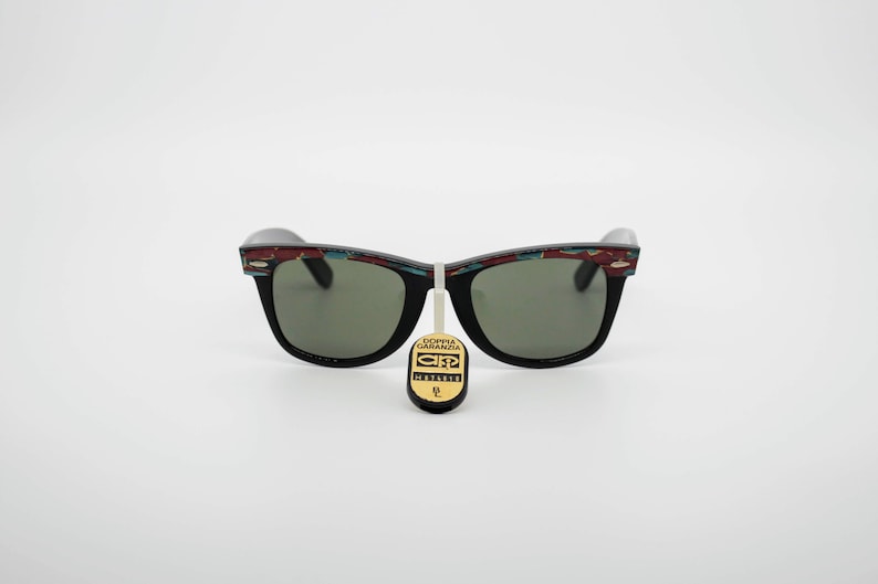 May include: Black sunglasses with green lenses and a decorative pattern on the top of the frames. The pattern includes red, blue, and white geometric shapes. A tag with text hangs from the sunglasses.