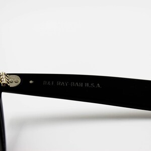 May include: Close-up of the black frame of a pair of sunglasses with brown lenses. The sunglasses have a metal hinge and the text "B&L RAY-BAN U.S.A." is printed on the frame.