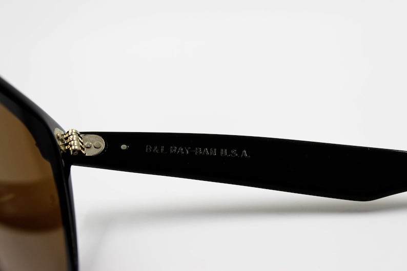 May include: Close-up of the black frame of a pair of sunglasses with brown lenses. The sunglasses have a metal hinge and the text "B&L RAY-BAN U.S.A." is printed on the frame.