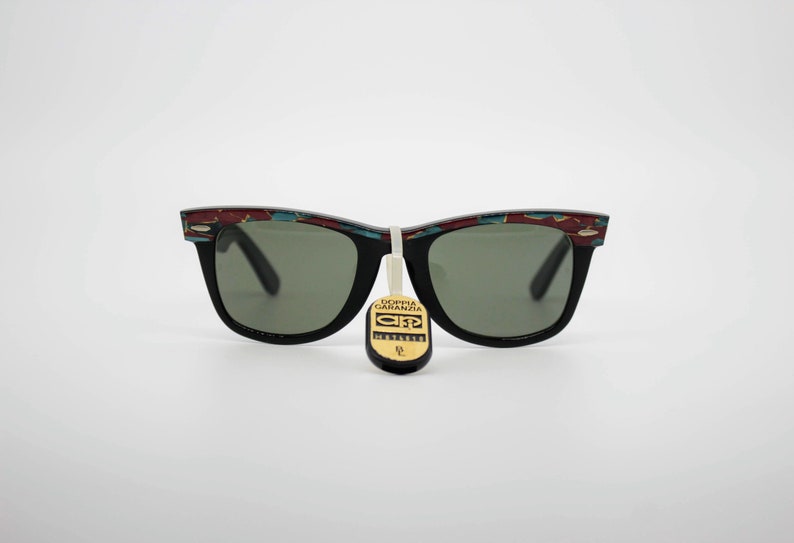 May include: Black sunglasses with a decorative pattern on the top of the frames. The frames have a rectangular shape with green lenses. A tag hangs from the sunglasses with text.