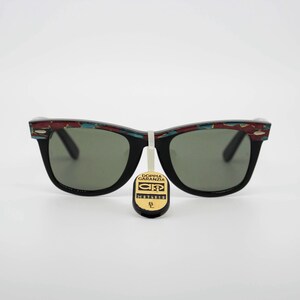 May include: Black sunglasses with a decorative pattern on the top of the frames. The lenses are a dark green color. A tag with text hangs from the center of the sunglasses.