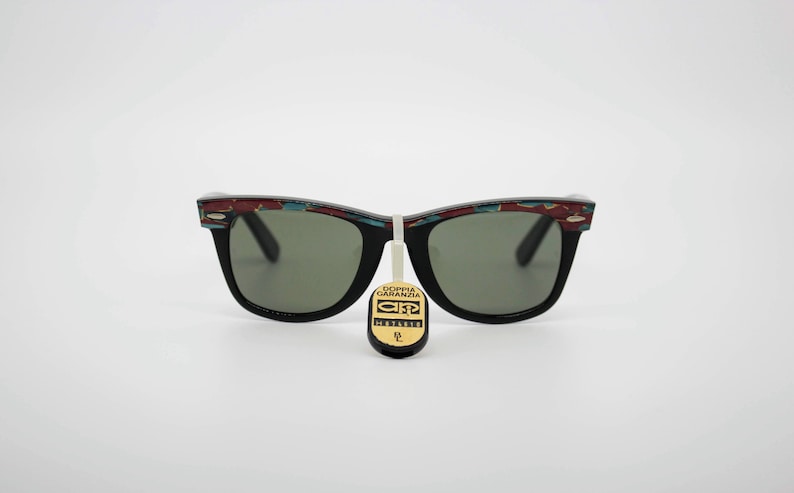 May include: Black sunglasses with a decorative pattern on the top of the frames. The lenses are a dark green color. A tag with text hangs from the center of the sunglasses.