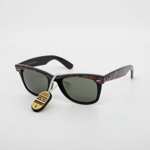May include: Black sunglasses with a patterned design on the frames. The sunglasses have dark lenses and a tag attached. The frames have a mix of red, blue, and white colors. The sunglasses are a classic style.