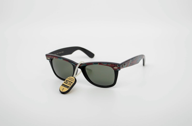 May include: Black sunglasses with a patterned design on the frames. The sunglasses have dark lenses and a tag attached. The frames have a mix of red, blue, and white colors. The sunglasses are a classic style.