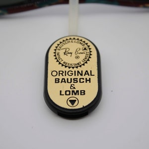 May include: Close-up of a black and gold Ray-Ban tag. The tag has the Ray-Ban logo and the words "ORIGINAL BAUSCH & LOMB" printed on it. The tag is attached to a white string.