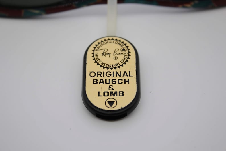 May include: Close-up of a black and gold Ray-Ban tag. The tag has the Ray-Ban logo and the words "ORIGINAL BAUSCH & LOMB" printed on it. The tag is attached to a white string.