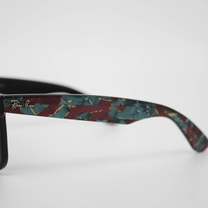 May include: Black Ray-Ban sunglasses with a patterned design in teal, burgundy, and gold. The Ray-Ban logo is visible on the temple. The sunglasses have dark lenses and a classic frame shape.