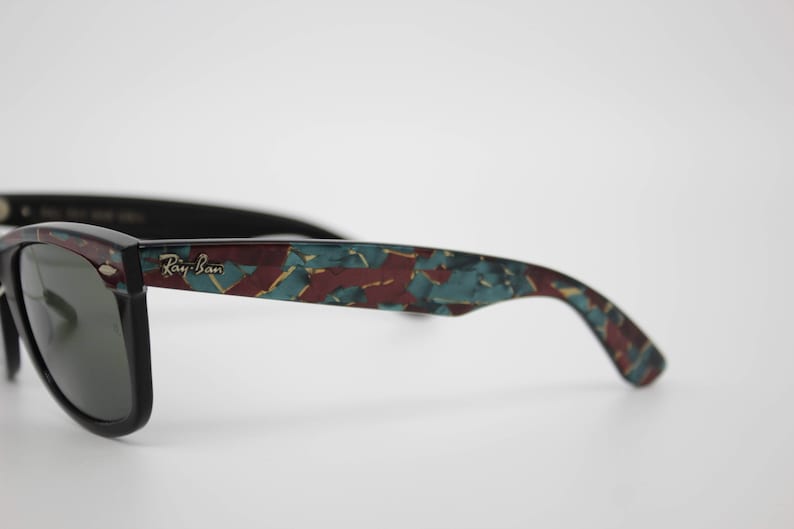 May include: Black Ray-Ban sunglasses with a patterned design in teal, burgundy, and gold. The Ray-Ban logo is visible on the temple. The sunglasses have dark lenses and a classic frame shape.