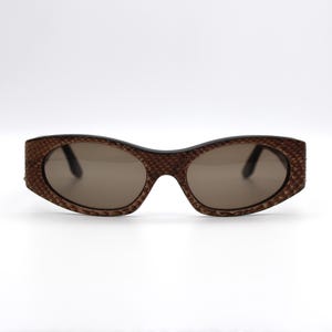 Könnte beinhalten: Braune ovale Sonnenbrille mit strukturiertem Muster auf den Rahmen. Die Gläser sind in einem gedämpften Braunton gehalten. Die Sonnenbrille ist vor einem weißen Hintergrund platziert.