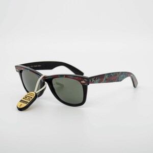 May include: Black sunglasses with a patterned design on the arms. The sunglasses have a tag attached with text. The lenses are dark, and the frames are black. The arms have a pattern of red, blue, and green.