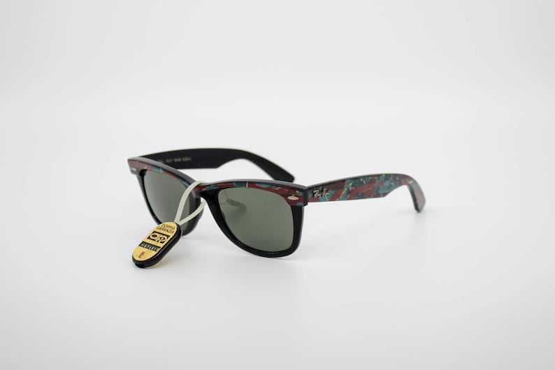 May include: Black sunglasses with a patterned design on the arms. The sunglasses have a tag attached with text. The lenses are dark, and the frames are black. The arms have a pattern of red, blue, and green.