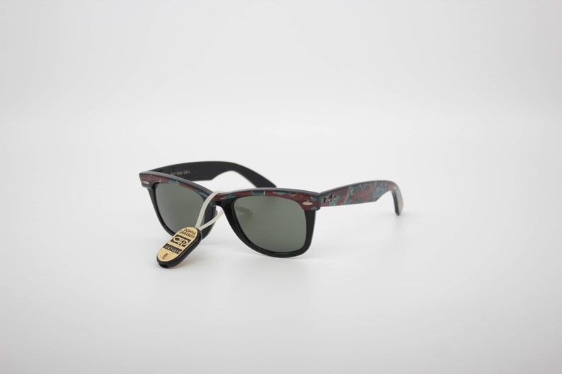 May include: Black sunglasses with a patterned design on the frames. The frames have a mix of red, teal, and black colors. The sunglasses have a tag attached with text. The lenses are dark gray.