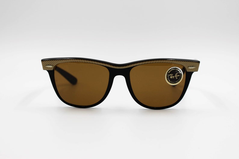 May include: Black sunglasses with brown lenses and a gold-colored frame. The frame has a textured design and the sunglasses have a Ray-Ban logo sticker on the right lens.