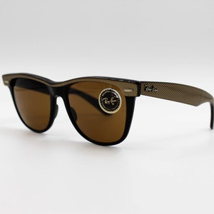 May include: Brown sunglasses with a black frame and gold accents. The sunglasses have a Ray-Ban logo on the side.
