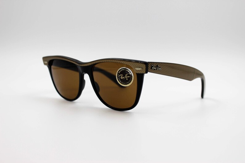 May include: Brown sunglasses with a black frame and gold accents. The sunglasses have a Ray-Ban logo on the side.