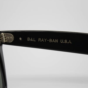 May include: Close-up of black sunglasses with the text "B&L RAY-BAN U.S.A." printed in gold on the temple. The sunglasses have a classic design with a rectangular frame and dark lenses. The hinge is silver.