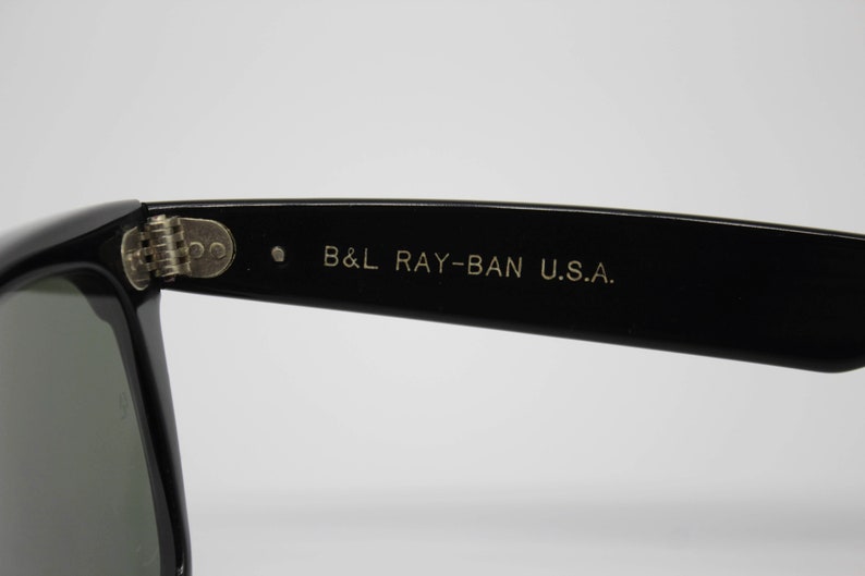 May include: Close-up of black sunglasses with the text "B&L RAY-BAN U.S.A." printed in gold on the temple. The sunglasses have a classic design with a rectangular frame and dark lenses. The hinge is silver.