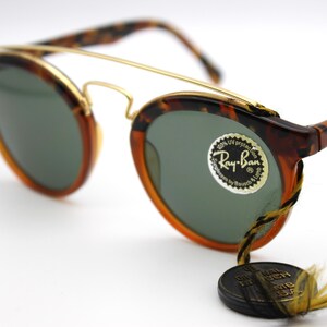 May include: Tortoise shell Ray-Ban sunglasses with gold accents and green lenses. The sunglasses have a round shape and a classic design. The Ray-Ban logo is visible on the right lens.