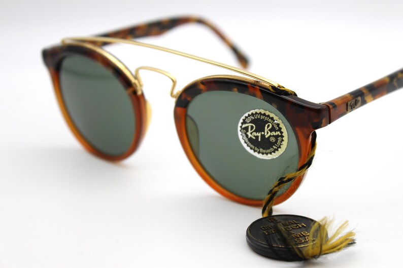 May include: Tortoise shell Ray-Ban sunglasses with gold accents and green lenses. The sunglasses have a round shape and a classic design. The Ray-Ban logo is visible on the right lens.