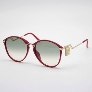May include: Red round sunglasses with gold metal arms and a gold accent on the side. The lenses are a gradient of gray.