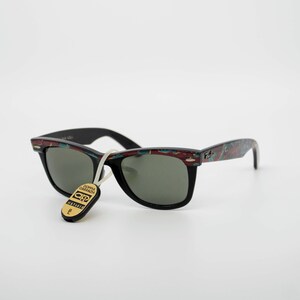 May include: Black sunglasses with a patterned design on the arms and a tag attached. The sunglasses have a classic shape with dark lenses. The arms feature a mix of red, blue, and green colors. The tag has gold lettering.