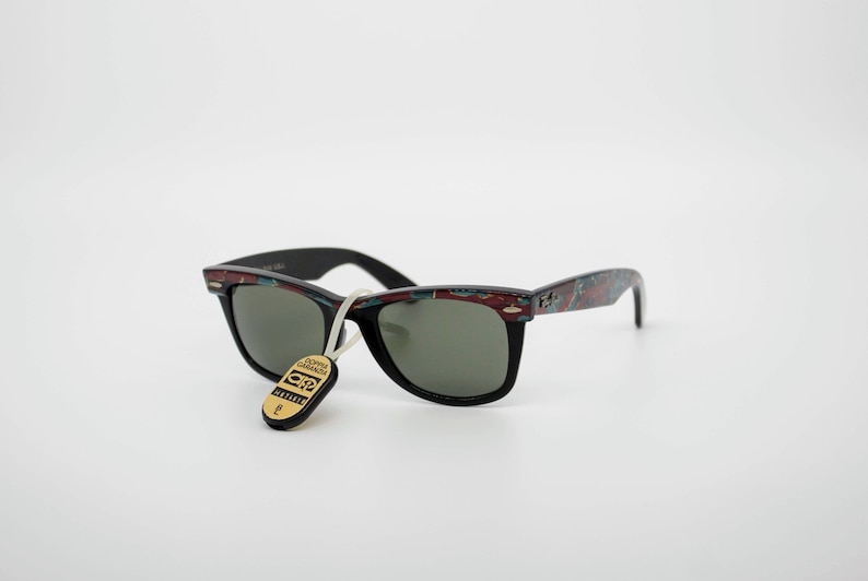 May include: Black sunglasses with a patterned design on the arms and a tag attached. The sunglasses have a classic shape with dark lenses. The arms feature a mix of red, blue, and green colors. The tag has gold lettering.