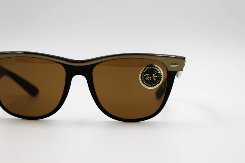 May include: Brown Ray-Ban sunglasses with a gold-colored frame and a gold sticker that says "Ray-Ban" on the right lens.