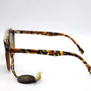 May include: Tortoise shell sunglasses with gold accents and a brown leather strap. The sunglasses have a unique design with a curved frame and a wide lens.