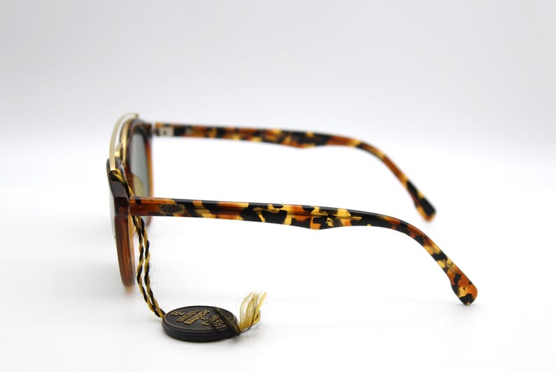 May include: Tortoise shell sunglasses with gold accents and a brown leather strap. The sunglasses have a unique design with a curved frame and a wide lens.