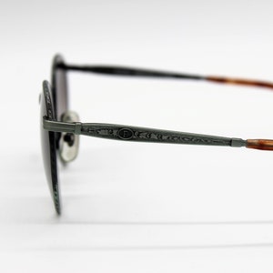 Op de afbeelding: Een paar ronde, donkergrijze zonnebrillen met een zilveren metalen montuur en bruine schildpadpootjes. Het montuur heeft een gegraveerd ontwerp.