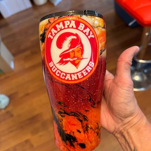 May include: A tumbler featuring the Tampa Bay Buccaneers logo in a red and white circle. The tumbler has a glittery red and orange design with black accents. The logo includes a pirate with a feathered hat and the words "TAMPA BAY BUCCANEERS".