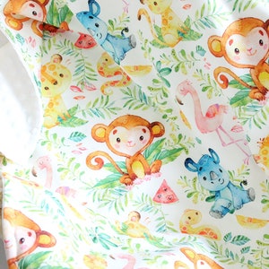 May include: A white fabric baby blanket with a jungle animal print. The print features monkeys, giraffes, rhinos, flamingos, and watermelon slices. The animals are illustrated in a watercolor style.