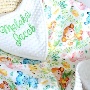Op de afbeelding: Wit babydekentje met een jungle dierenprint met apen, neushoorns, giraffen en ananas. Het dekentje heeft een witte minky achterkant met de naam "Malakai Jacob" geborduurd in groen.
