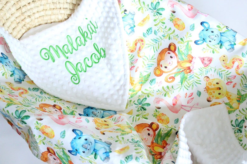 Op de afbeelding: Wit babydekentje met een jungle dierenprint met apen, neushoorns, giraffen en ananas. Het dekentje heeft een witte minky achterkant met de naam "Malakai Jacob" geborduurd in groen.