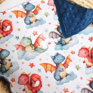 May include: A white baby blanket with a blue minky backing. The blanket features a repeating pattern of watercolor dragons with stars and clouds. The dragons are in shades of blue, green, and red.