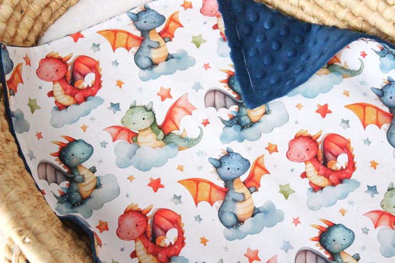 May include: A white baby blanket with a blue minky backing. The blanket features a repeating pattern of watercolor dragons with stars and clouds. The dragons are in shades of blue, green, and red.