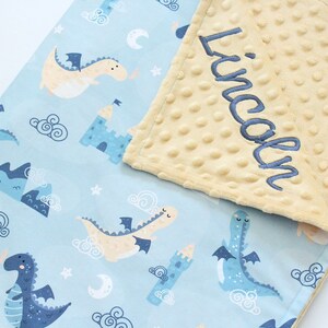 May include: A light blue baby blanket featuring a pattern of dragons, castles, clouds, stars, and moons. One side is smooth fabric, while the other is a textured, pale yellow minky fabric with the name Lincoln embroidered in dark blue script.